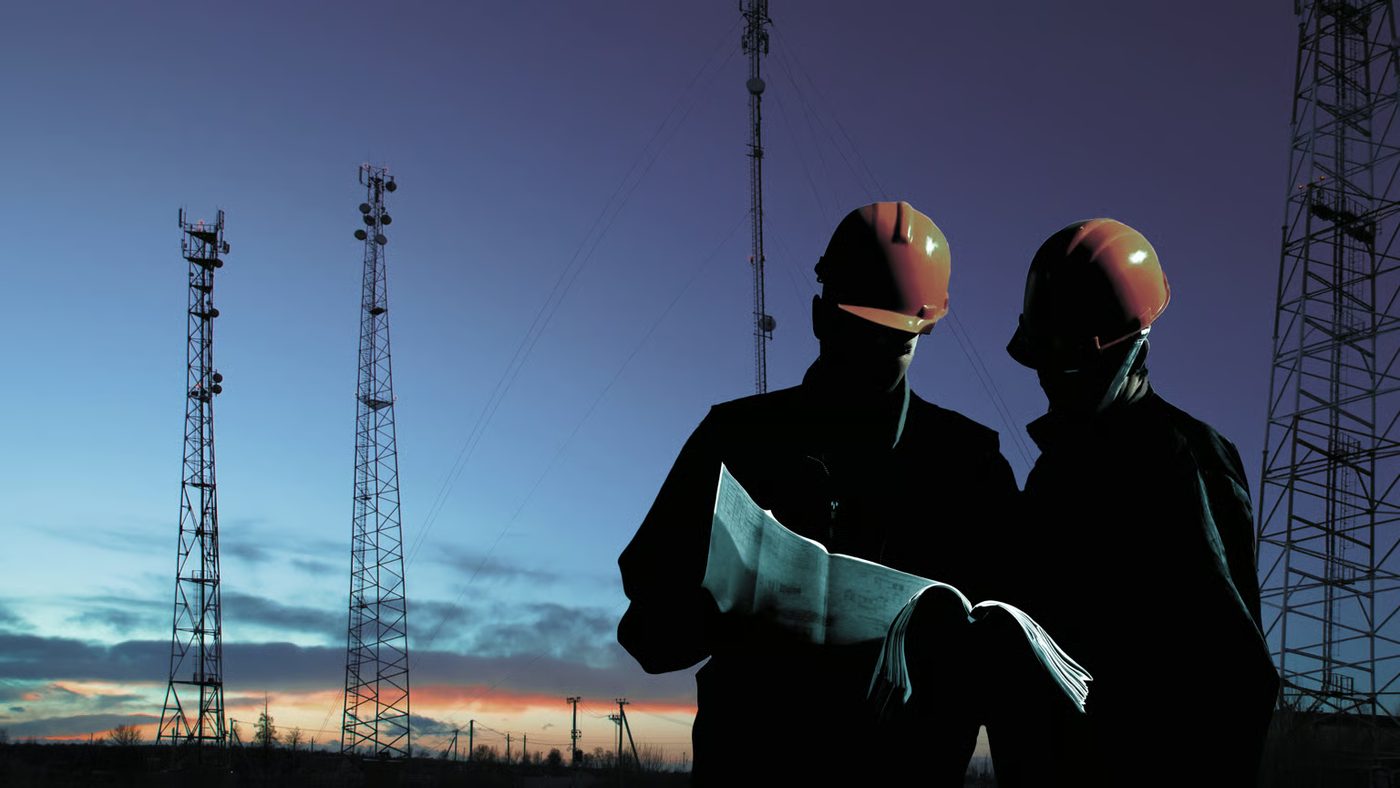 Workers silhouetted against the sky reviewing plans next to wireless towers at sunset.